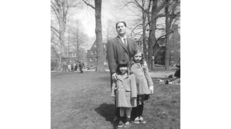 1966? Black-and-white vintage outdoor photo of a man wearing an overcoat standing on a grassy lawn with college buildings in the background. His hands are on the shoulders of two small girls in front of him: a smiling hark-haired girl with bangs and a shy-looking girl. Her hair looks light brown but it's actually red in real life.
