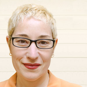 Nicola Ginzler, a white woman with short blond hair and black-framed glasses, in a peach-colored blouse.