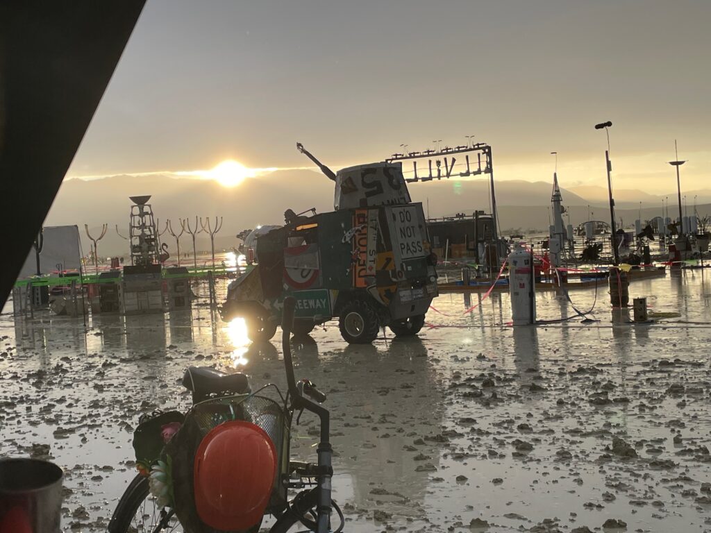 Looking from the Esplanade towards the playa after one bout of rain, the playa is soaked. A "mutant vehicle" is in front of the iron Illumination Village "Ill Ville" gate. The sun is setting and the light is reflected in the wet ground.