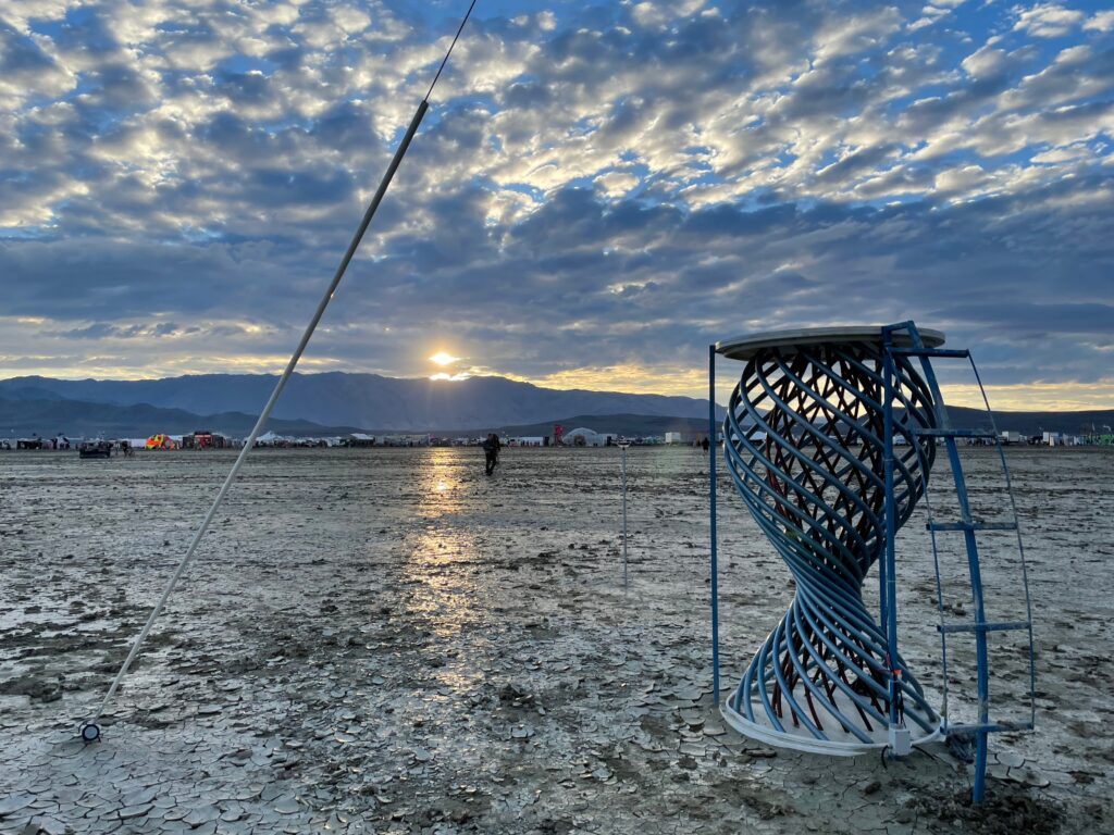 In the foreground, a structure of a black helix inside a blue helix made out of bent steel pipe. Surrounded by wet playa. in the far distance, the sun is low over black mountains. High rain clouds.