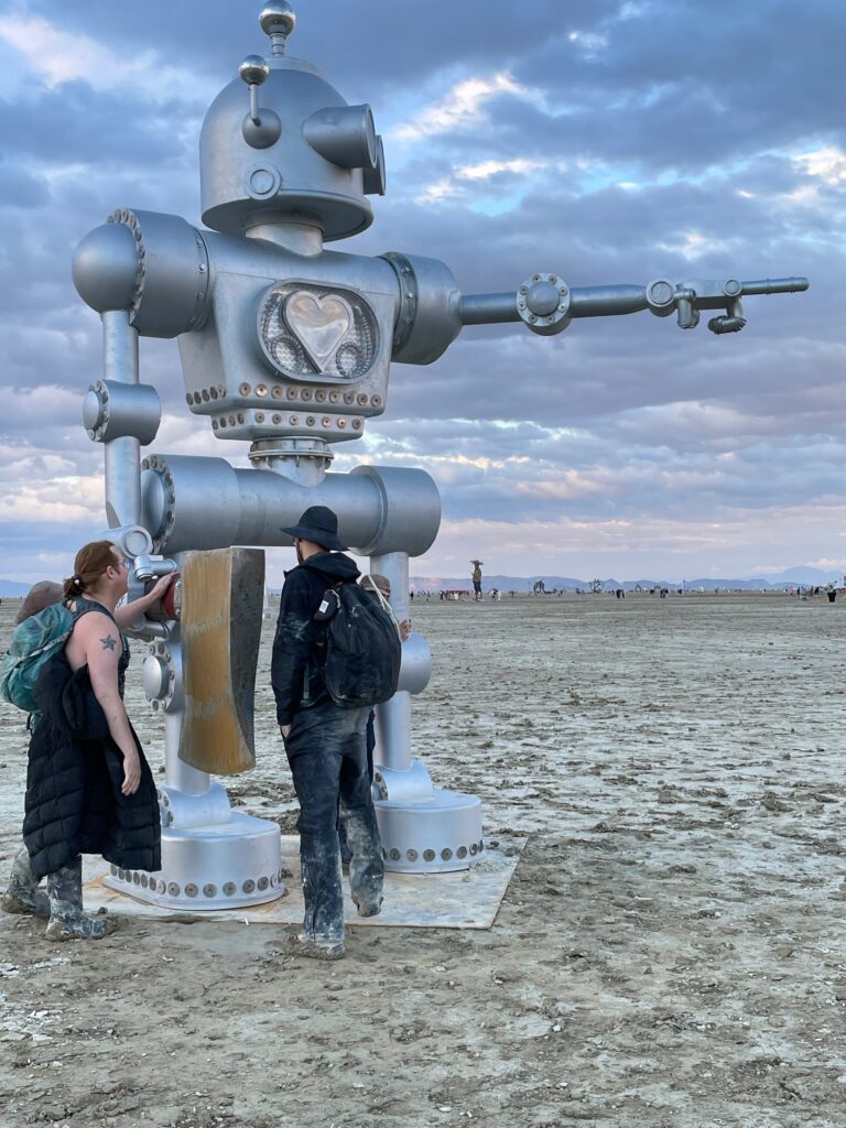 Two of my FLG crew mates stand in front of a 15' high silver robot sculpture. The robot is pointing with its left hand and holds a massive wooden axe in its right hand.