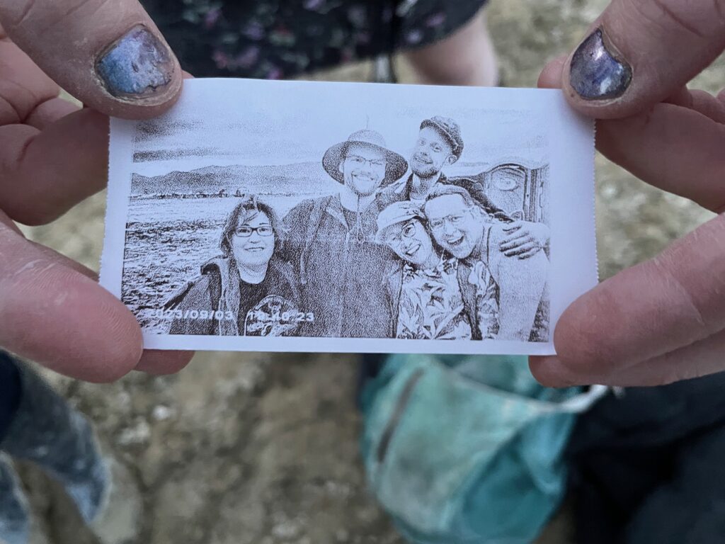 Two hands with worn-off nail polish hold a scrap of thermographic paper with the images of 5 smiling people on it.