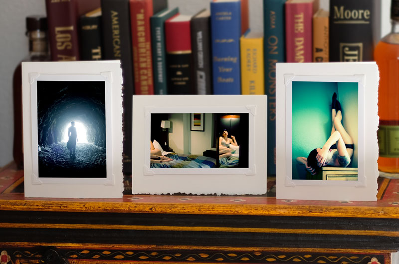 Photo of 3 greeting cards on a bookcase, standing up, each with a photo attached to it with photo corners. Left card: dark blue tunnel with slender female figure backlit against the entrance. Center: matter-of-fact woman in lingerie on hotel bed, part of her body and face reflected in a mirror. Cool-toned bedspread with warm-toned skin. Right: woman lying on her back on a dresser in the corner of a room. She's lying on her back with her legs up the wall. Blue and green tones with pale skin.