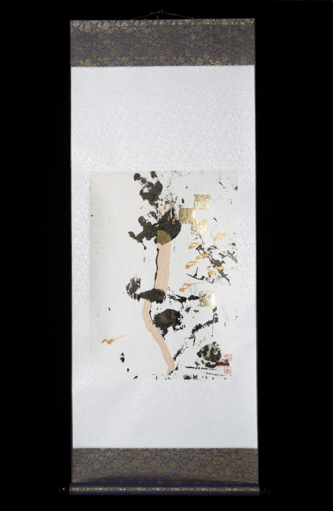 Evoco performance/artwork #4: pale cream brocade fabric scroll with paper mounted on it. Paper has seemingly random black paint impressions. Scattered smears have been added in gold paint; squares have been added in gold leaf.