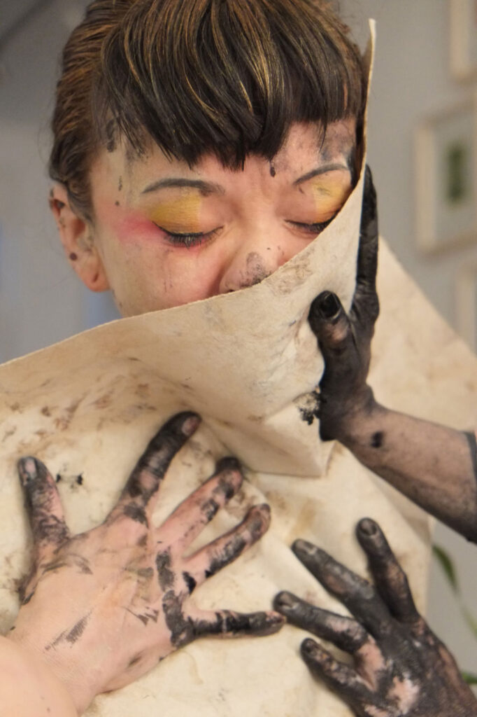 Evoco performance/artwork#2: three hands covered in paint pressing soft paper against woman's cheek