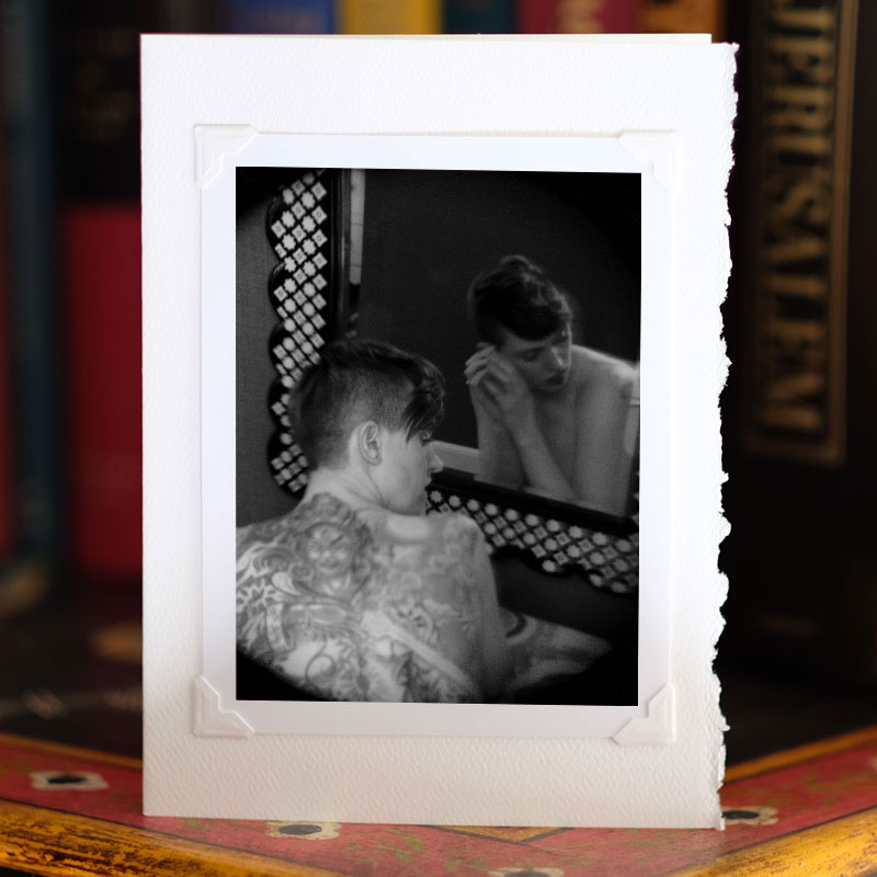 Photo of greeting card on a bookcase, standing up, with a photo attached to it with photo corners. Black and white image of a woman with her back to the viewer, looking in a mirror. She has a large elaborate tattoo of a demon on her back. Her face is visible in the mirror.