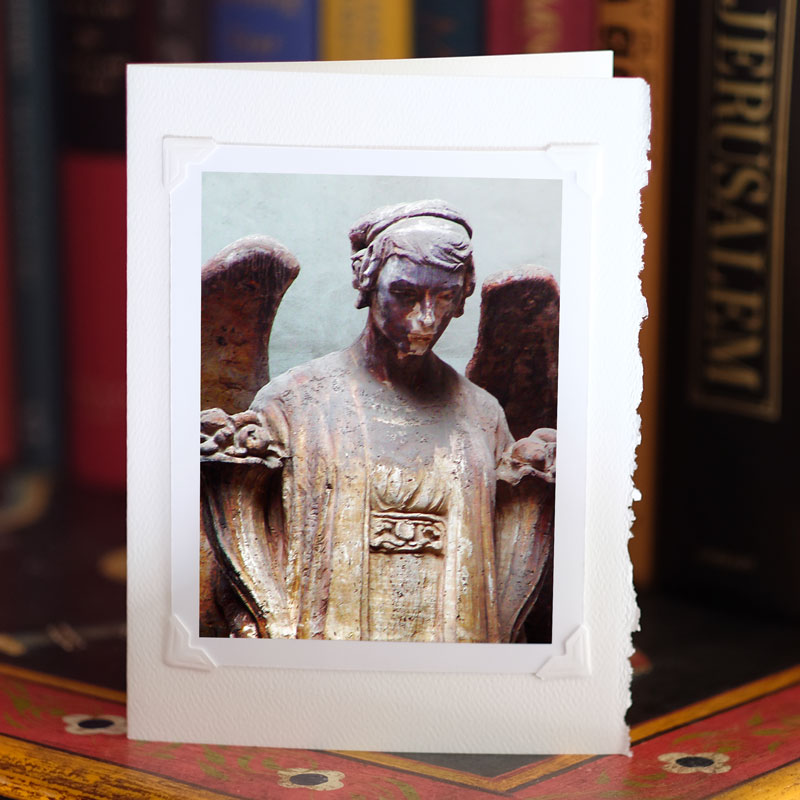 Photo of greeting card on a bookcase, standing up, with a photo attached to it with photo corners. Color image: a statue of a winged graveyard angel from the waist up.