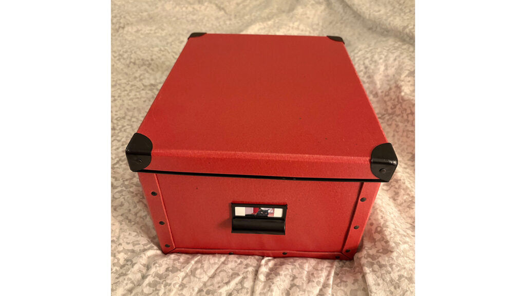 Photo of a red heavy cardboard box with black corners on the lid, on a light gray patterned comforter. A label on the front of the box has a photo of a black cat lying on a flowered beige comforter.