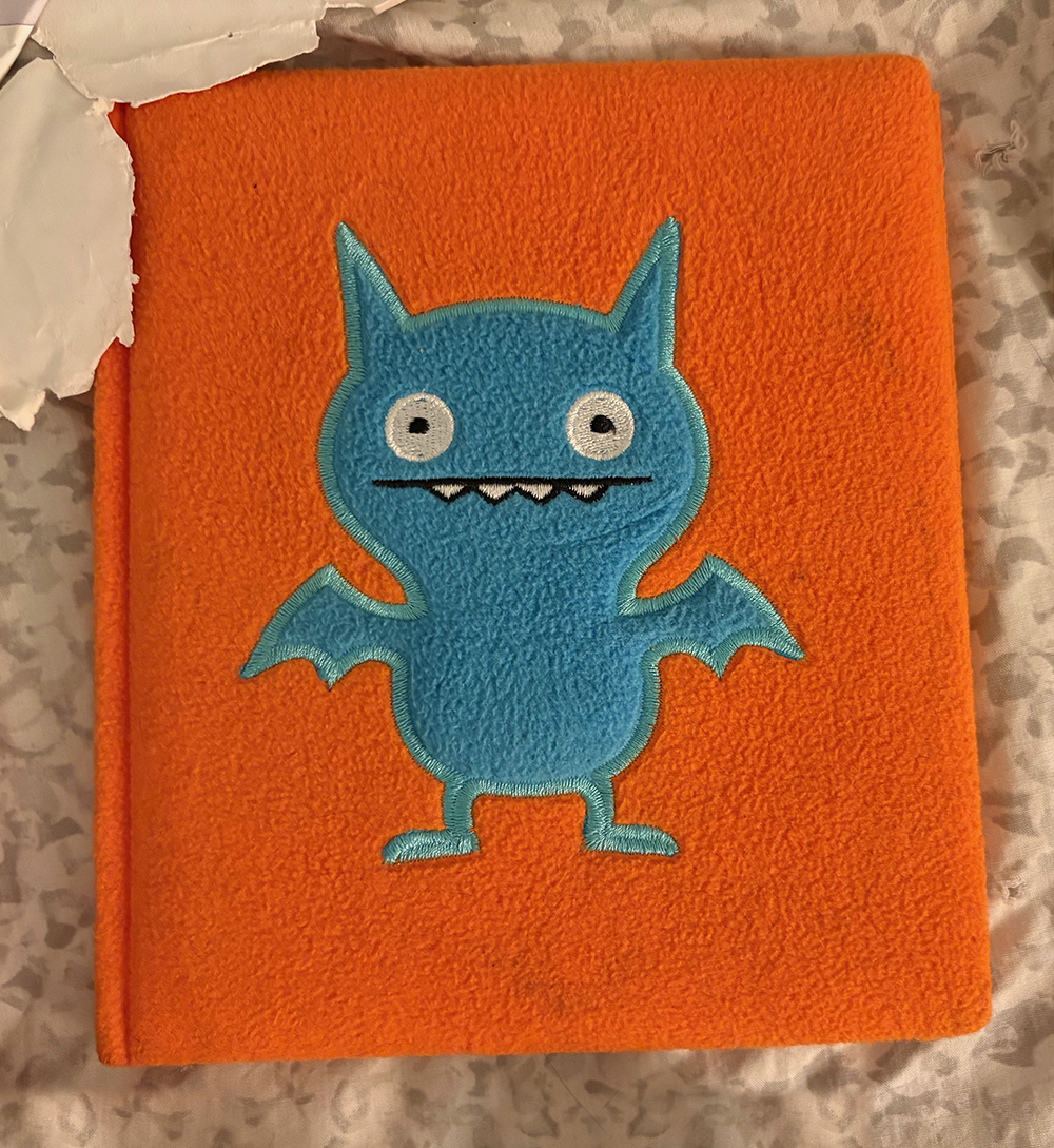 Photo of the orange felt cover of a journal with a blue stylized "Ice Bat" figure in turquoise blue felt, on a light gray comforter. Ice Bat is one of the UglyDolls plush toys. 