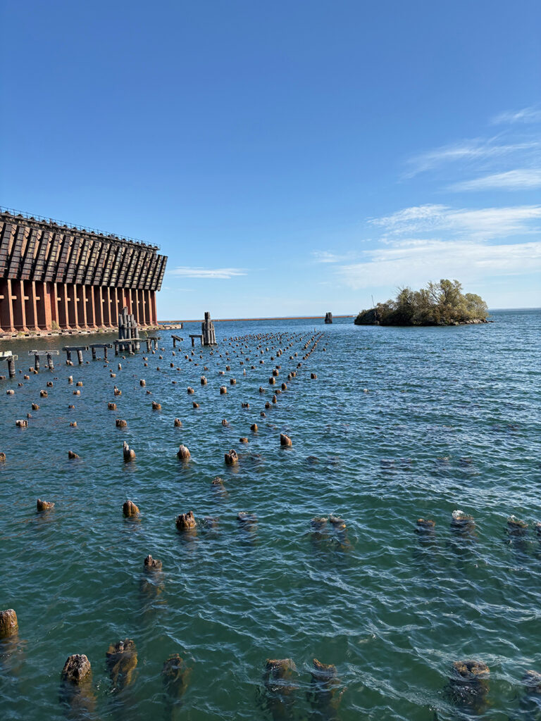 On the right, in foreground, stumps of wooden piles for a former pier, in blue water. Huge concrete structure in left background.