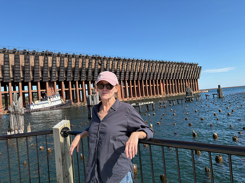 In foreground, woman with red hair leaning against an iron railing. In middle distance, stumps of wooden piles for a former pier, in blue water. Huge concrete structure in background.