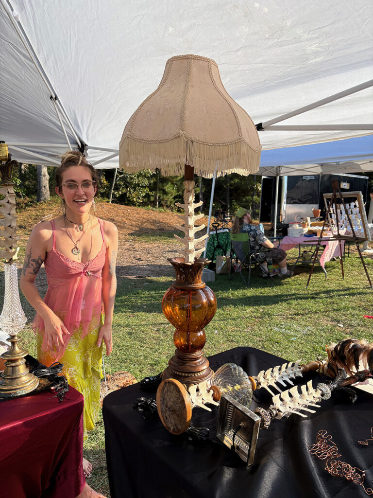 Smiling young woman under tent in open field with lamps on a table. Upright shafts of lamps have deer vertebrae threaded onto them and vintage fringed shades.