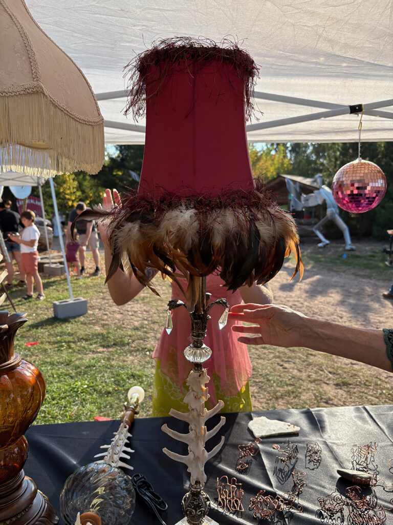 Lamp on table under tent in open field. Upright shaft of lamp has deer vertebrae threaded onto it. Vintage landshade is red fabric with a fronge of feathers added around the bottom.