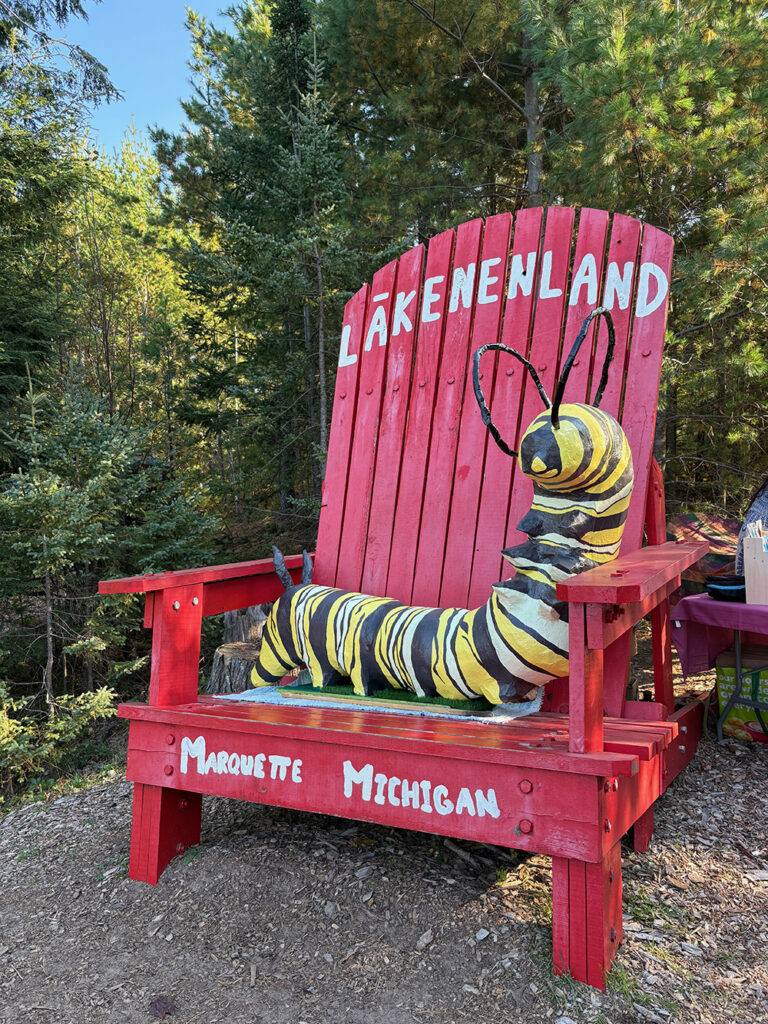 Huge Adirondack-style chair, painted red, with a large, fanciful yellow and black caterpillar on the seat and back. The the top of the back says "LAKENENLAND" in white type; the front of the seat says "MARQUETTE MICHIGAN."