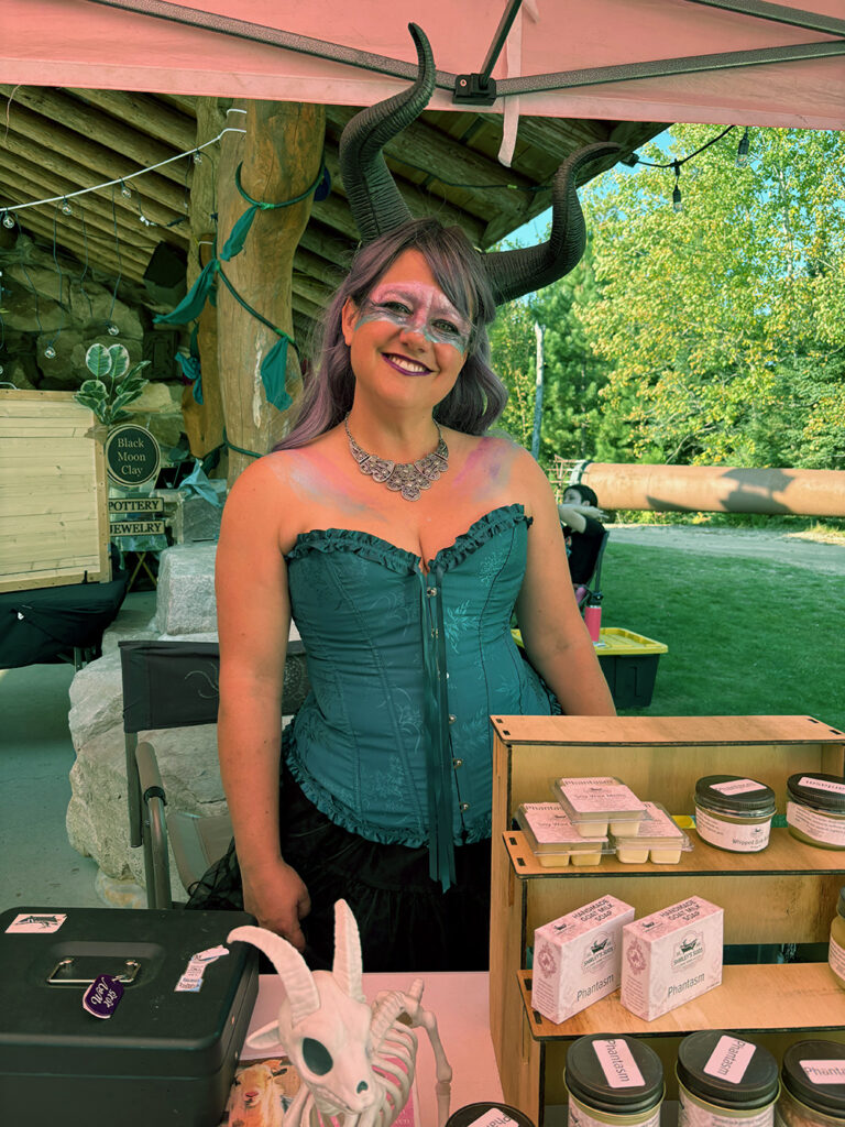Smiling woman with devil horns and teal corset, under a tent in an open field. In front of her is a product display of soaps and candles.
