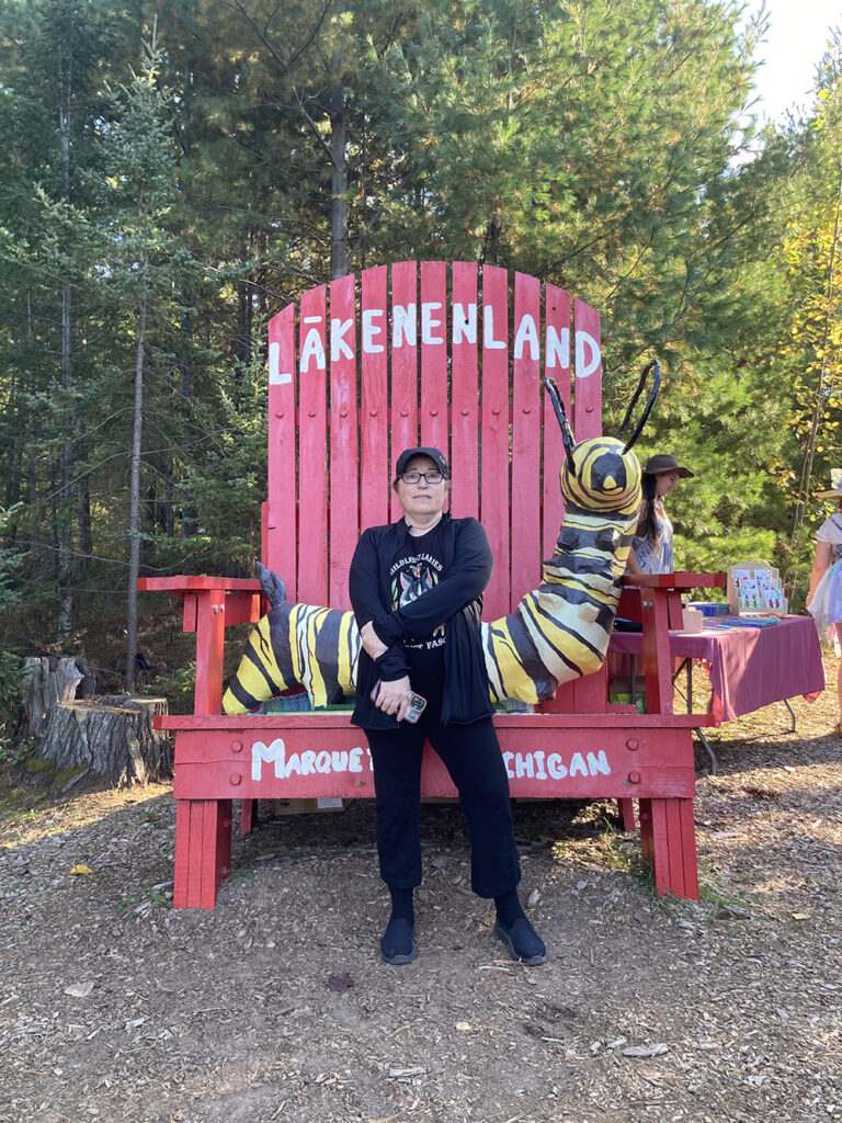 Big red chair with woman with black hat standing in front center, neutral expression.