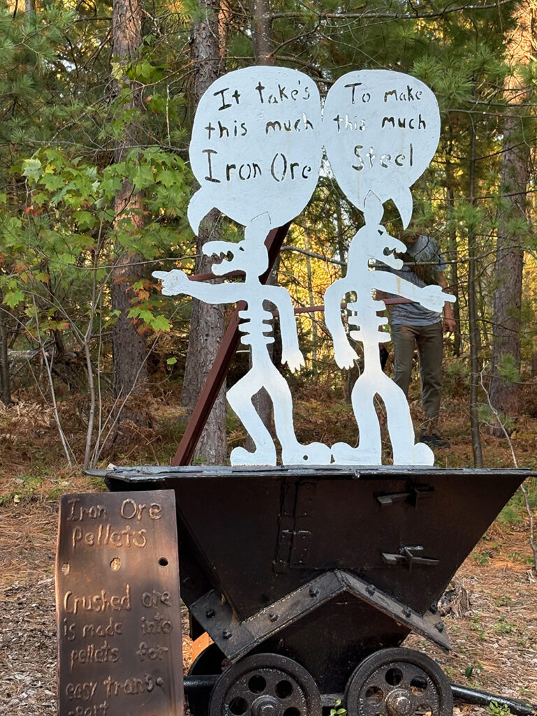 Two fanciful metal figures pointing in opposite directions, with a speech bubble over each head. Left: "It takes this much Iron Ore" - right: "To make this much steel"