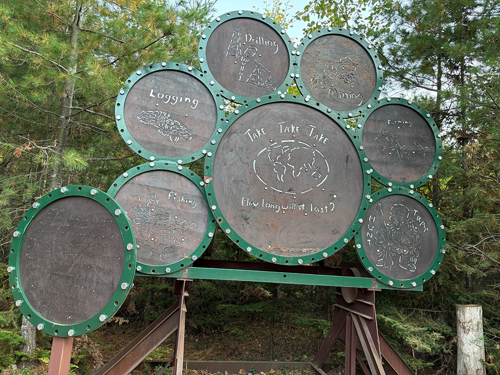 Metal sign: 1 big circle with 6 smaller circles around it, and 1 smaller circle off to the side. Cut into the circles are words and images. Big circle: "Take Take Take - How long will it last?" with graphic of globe map. Smaller circles, L-R, clockwise from bottom left: "Fishing," "Logging," "Drilling," "Mining," "Farming," "Trap - Hunt." At bottom left is another circle with small indecipherable type.