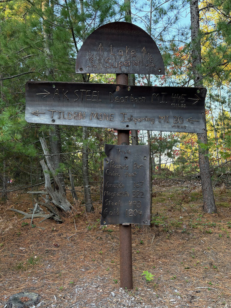 Upright sheets of metal with welds spelling out "Lake Superior - 1/2 mile" with forward arrows; "AK Steel - Dearborn, MI 445 miles," with right arrows; "Tilden Mine - Ishpeming, MI - 30 miles" with left arrows; "Trivia - Pounds per cubic foot - Granite 167- Magnetite 322 - Steel 493 - Gold 1204"