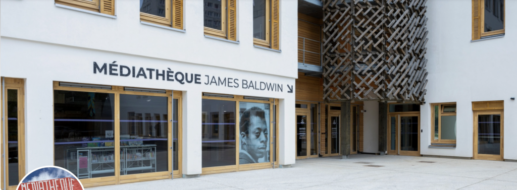 Exterior of the Mediatheque James Baldwin in Paris. Perspective shot of a light-colored building with large windows at ground level and the building name along its length. A duotone photo of James Baldwin fills the right-most window.