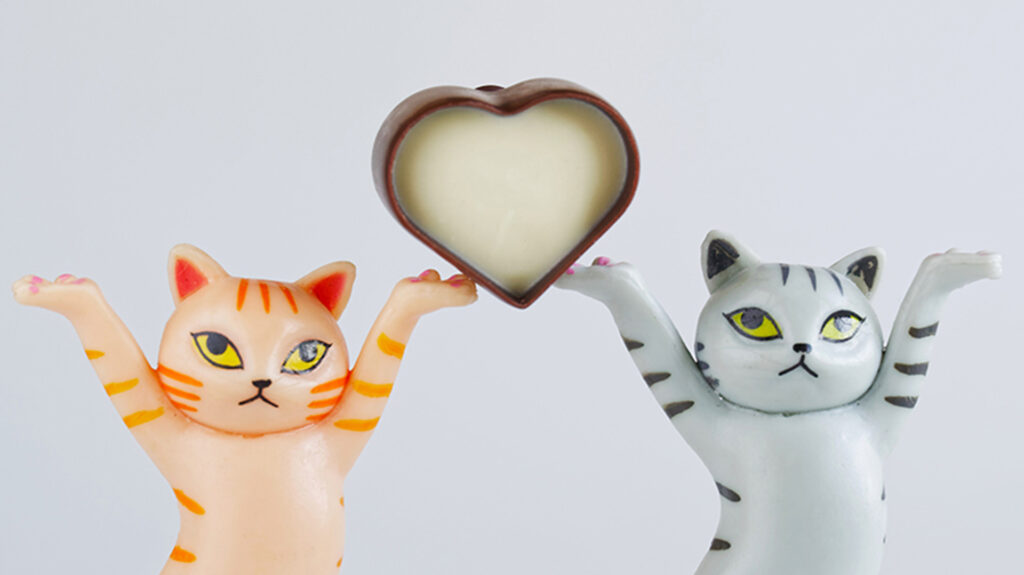 Cropped photo of toy kittens carrying a heart-shaped candy made of black and white chocolate. Pale blue background.