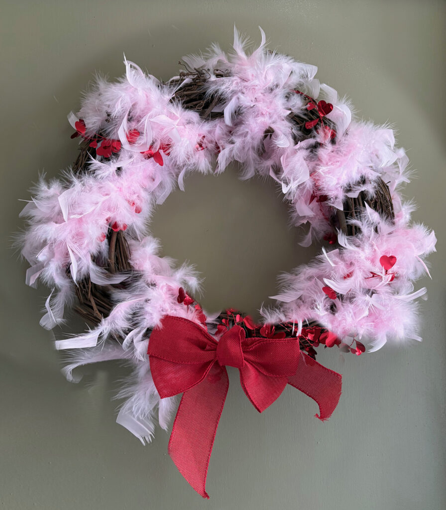 A wreath on a gray-green door. The wreath is a brown wicker circle with pale pink maribou feathers and a red metallic heart garland wound around it. There's a red ribbon bow at the bottom.
