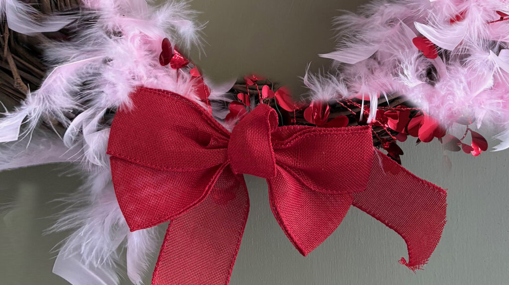 Tight crop on the ribbon of a wreath on a gray-green door. The wreath is a brown wicker circle with pale pink maribou feathers and a red metallic heart garland wound around it. There's a red ribbon bow at the bottom.