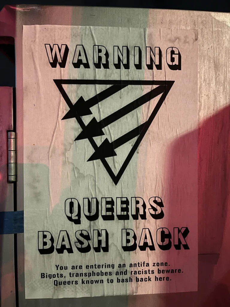 Photo of weathered vertical poster wheat-pasted to a wall. White background with black artwork and black, centered type. Top text in an all-caps, 3-D style stencil typeface: "WARNING." Next, a black outline of an upside-down equilateral triangle with three parallel black arrows through it, pointing down and to the left. Below that, in the same type style as above: "QUEERS BASH BACK." And at the bottom, in small stencil type: "You are entering an antifa zone. Bigots, transphobes and racists beware. Queers are known to bash back here."